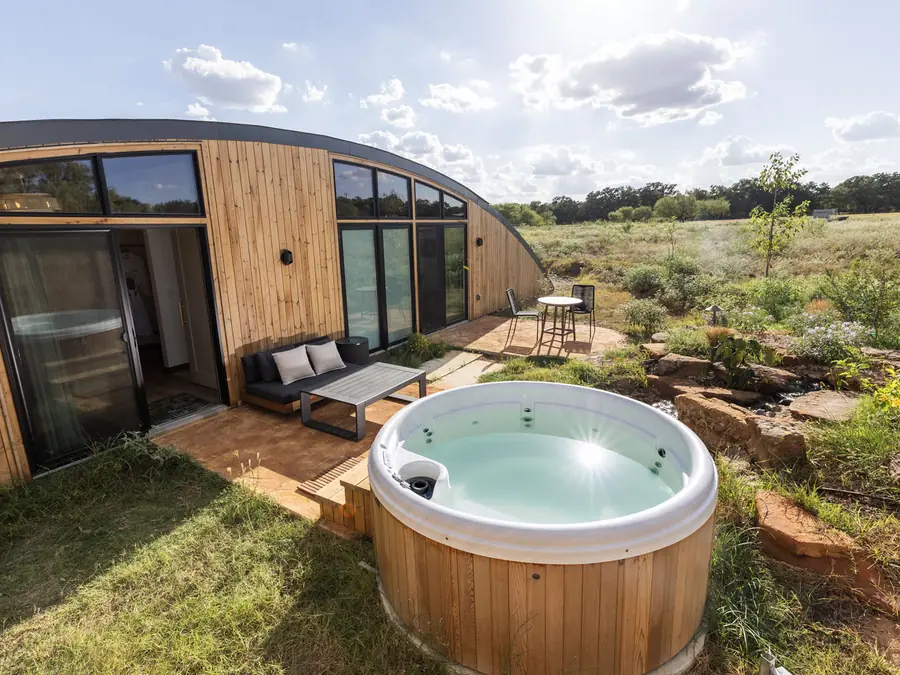 A curved timber-clad cabin with a deck, a round cedar hot tub and open meadow beyond