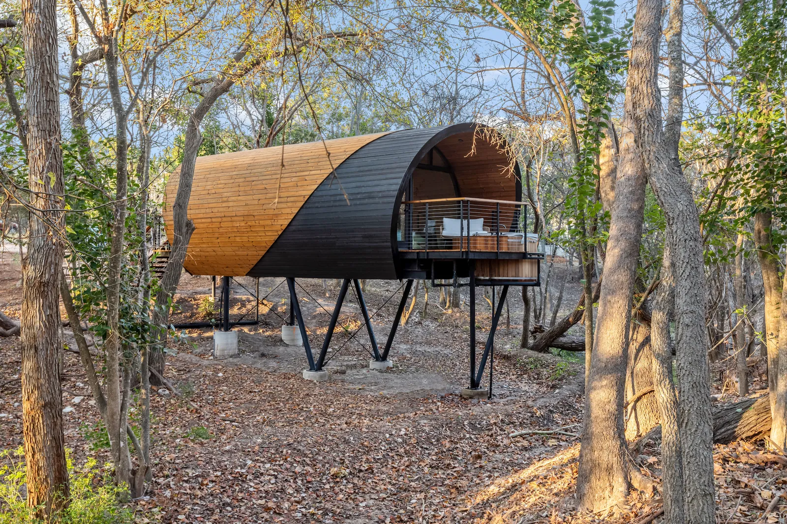 A barrel-vaulted cabin in timber and dark metal, raised on angled steel legs in a bare winter wood