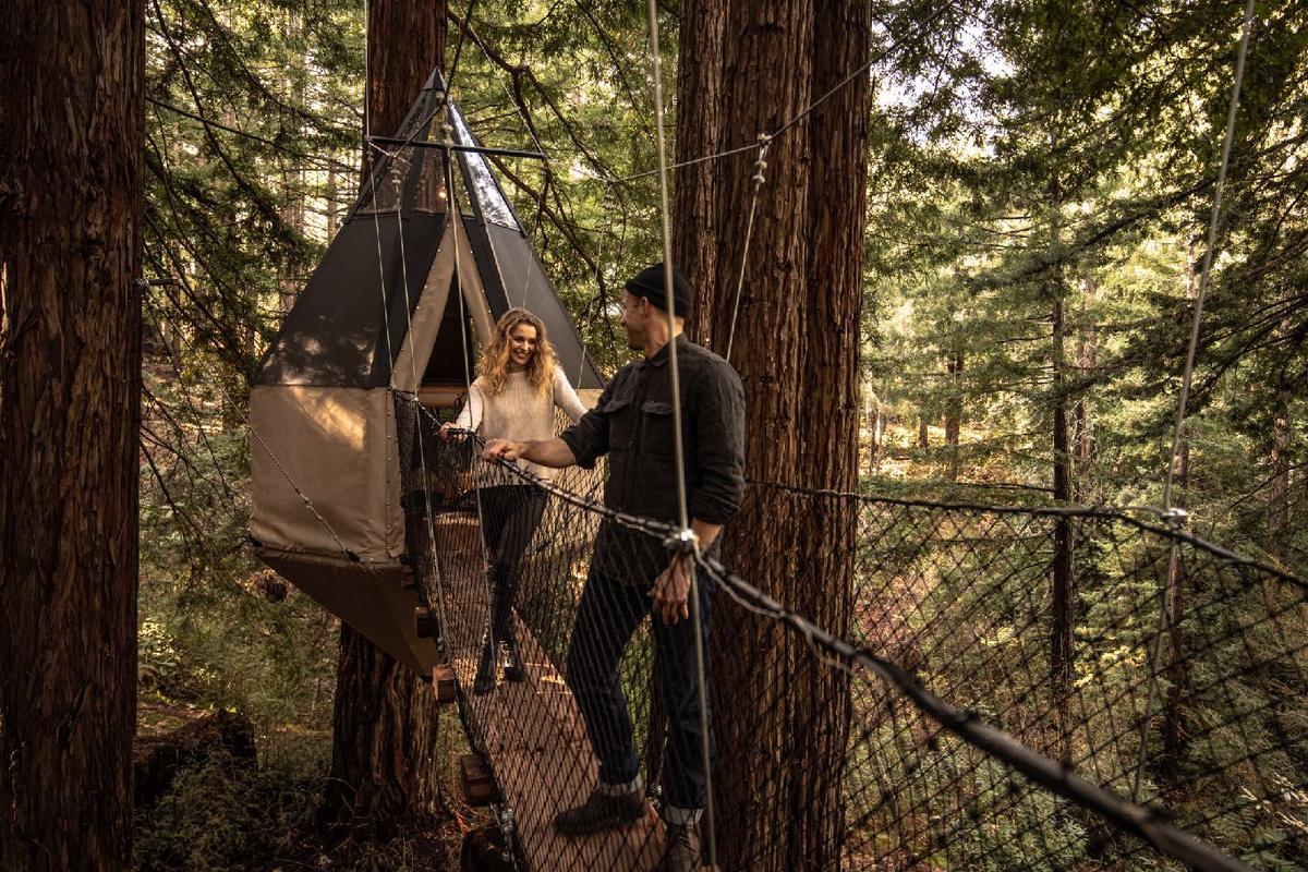 Two people on a rope suspension bridge beside a hanging canvas tent pod in the redwoods