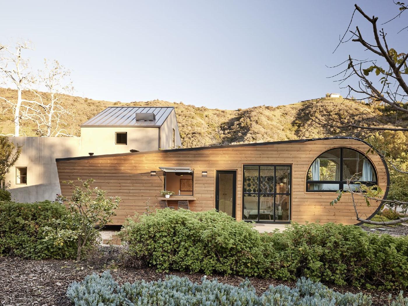 A low curved timber-clad house with a large round window, set into chaparral hills