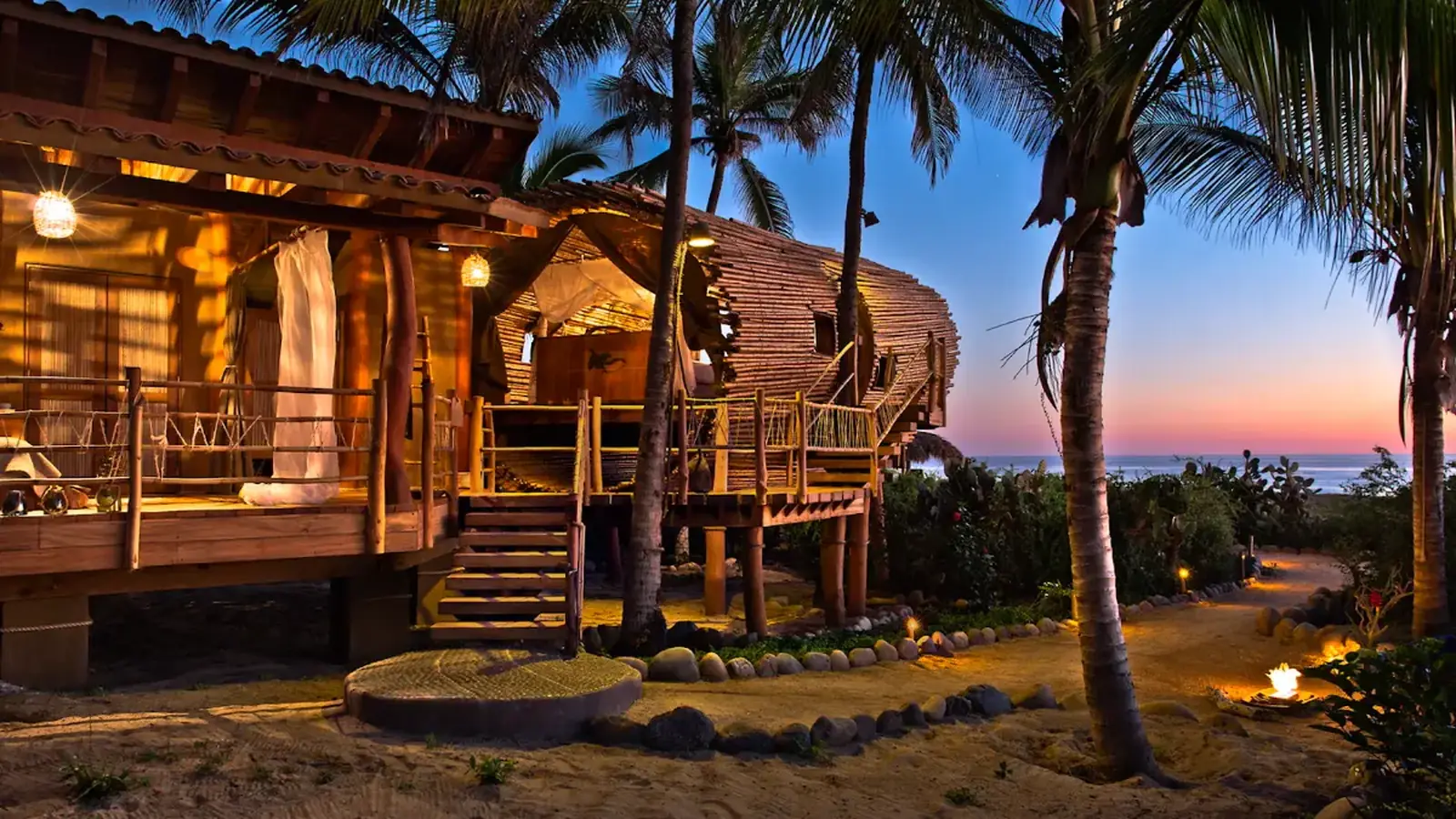 A timber treehouse suite among palms at dusk, warm light spilling from the deck