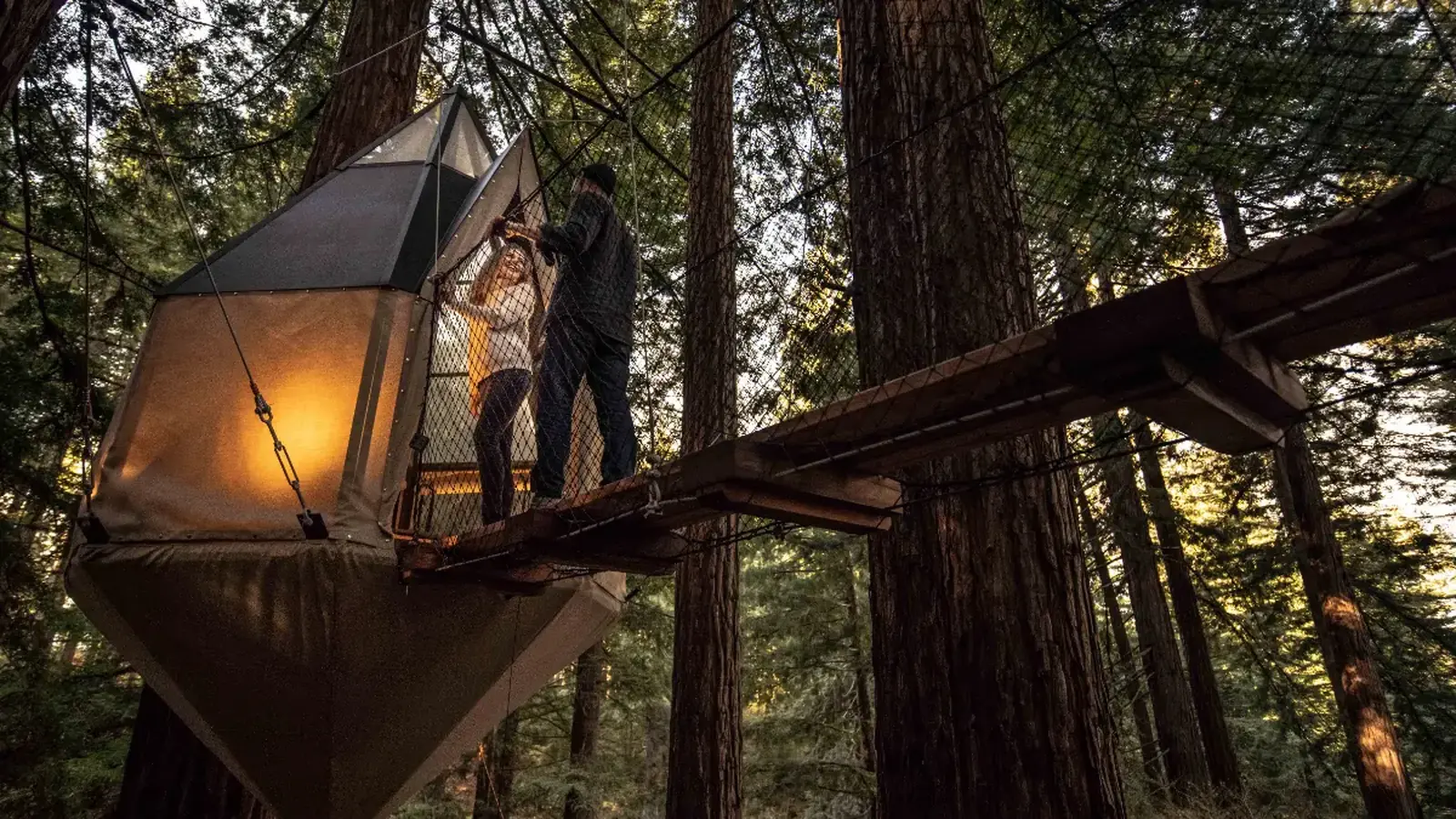 Two people crossing a lit rope bridge to an angular tent cabin high in the redwoods at night