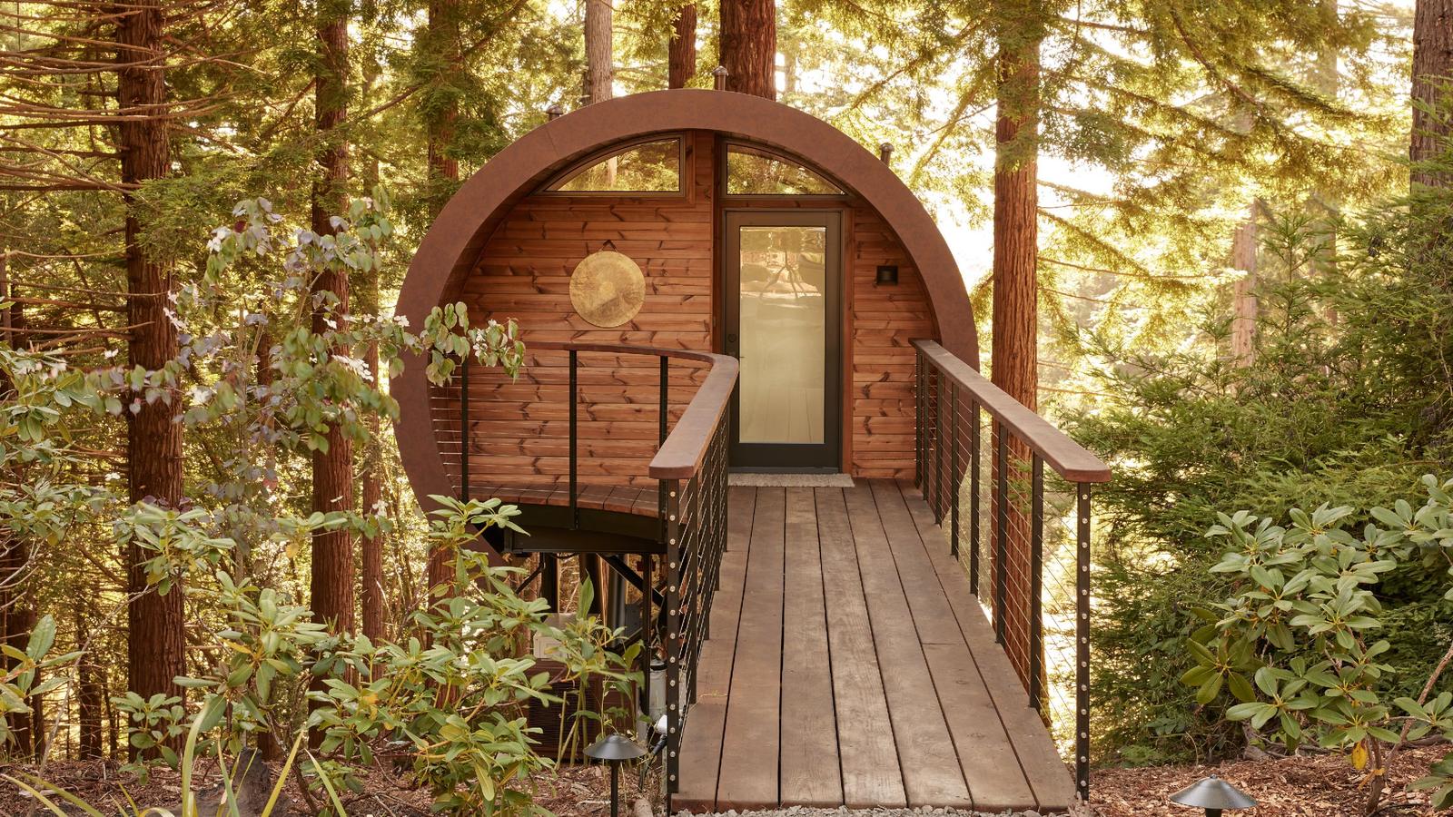A timber walkway leading to the round arched door of a barrel-vaulted treehouse