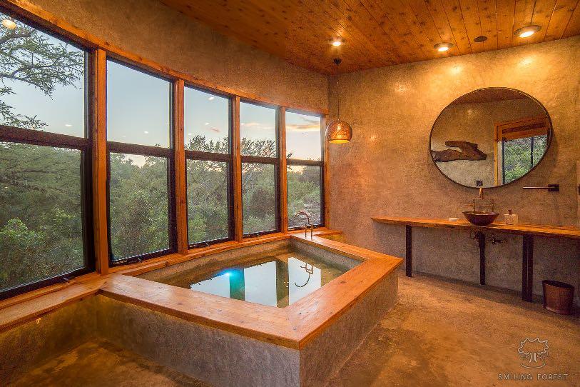 A timber interior with a wall of windows, a built-in soaking tub and a round mirror above a stone vanity